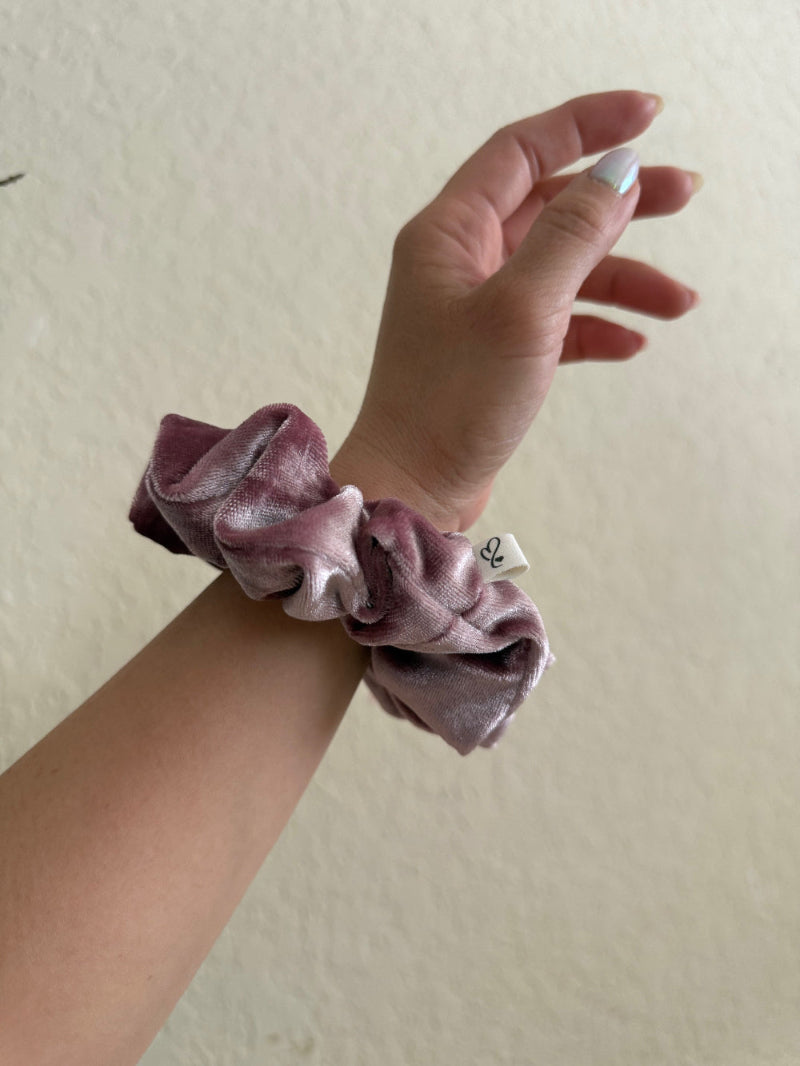 Hand wearing a pink scrunchie with a brand label on a plain background