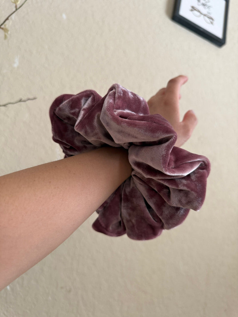 Hand holding a purple velvet scrunchie against a beige wall.