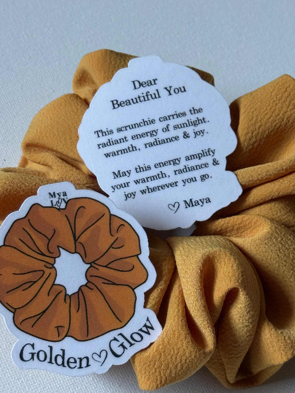 Brown scrunchie with a tag and 'Golden Glow' label on a light gray background