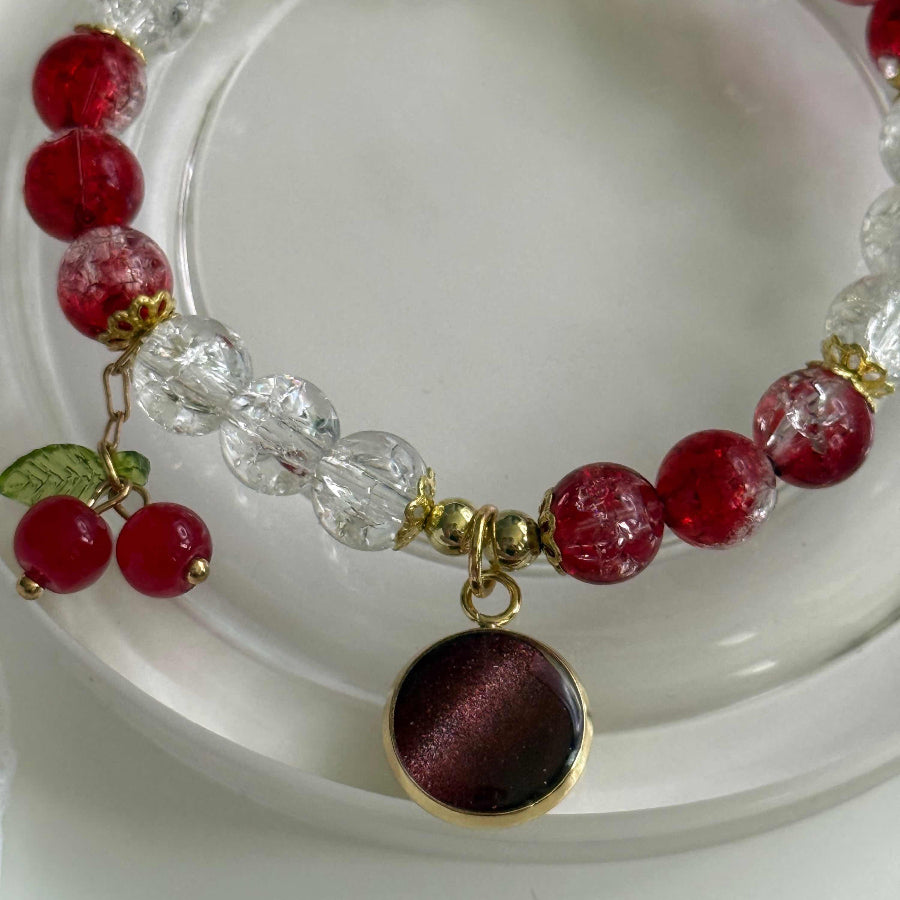 A beaded bracelet with red and clear faceted beads, gold-tone metal spacers, and a round dark red pendant with a gold border, displayed on a white surface.