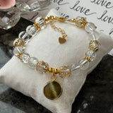 A beaded bracelet with clear faceted beads, gold-toned spacers, a small gold heart charm, and a larger round gold-toned pendant, displayed on a cream satin pillow.
