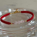 A red braided cord bracelet with three pale pink translucent beads and small gold spacer beads, displayed on a clear glass dish.