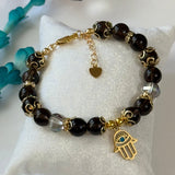 A beaded bracelet with dark brown and black beads, gold-toned metal spacers, a small clear faceted bead, a gold heart charm, and a gold Hamsa hand charm with a blue eye detail, displayed on a white textured pillow.