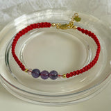 A red beaded bracelet with three purple beads and gold-tone clasp, displayed on a clear glass dish.