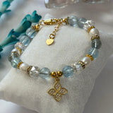 A beaded bracelet displayed on a white fabric cushion. The bracelet features light blue faceted beads, white pearl-like beads, clear crystal beads, and gold-tone metal accents, including a gold clover charm and a small gold heart charm.