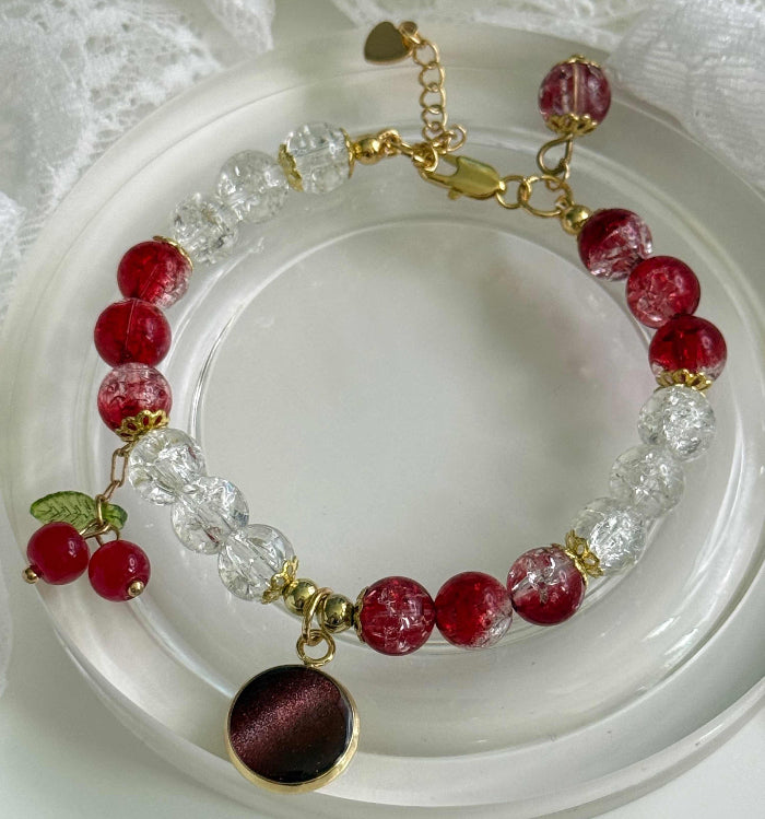 A beaded bracelet with red and clear faceted beads, gold-tone chain and connectors, cherry charms with green leaves, and a round dark red and gold pendant, displayed on a white dish.