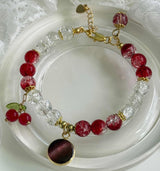 A beaded bracelet with red and clear faceted beads, gold-tone chain and connectors, cherry charms with green leaves, and a round dark red and gold pendant, displayed on a white dish.
