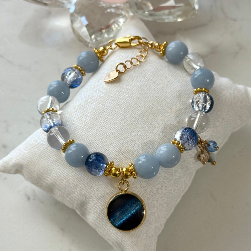 A beaded bracelet with light blue and dark blue beads, gold spacer rings, and a round dark blue cat-eye charm, displayed on a white fabric cushion.