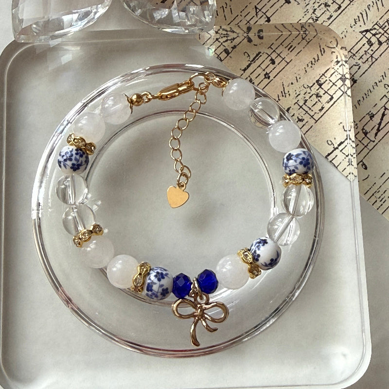 A beaded bracelet displayed on a clear round stand, featuring white and blue patterned beads, translucent clear beads, a small blue bead, gold-toned metal accents, and a dangling gold heart charm.