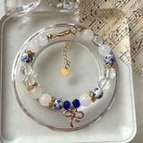 A beaded bracelet displayed on a clear round stand, featuring white and blue patterned beads, translucent clear beads, a small blue bead, gold-toned metal accents, and a dangling gold heart charm.