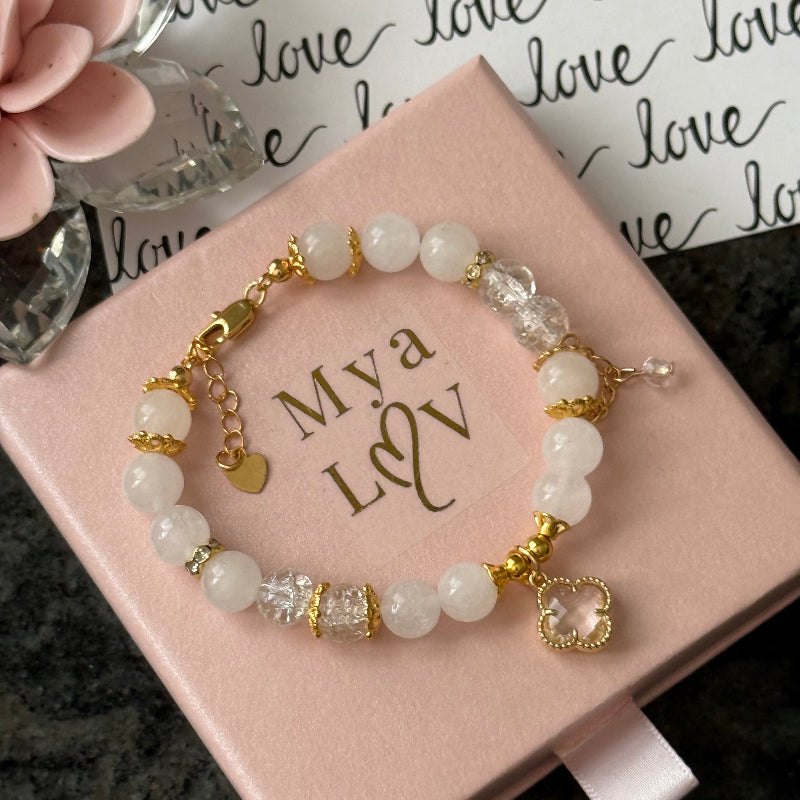 A beaded bracelet with white and clear beads, gold-toned spacer rings, a small heart charm, and a larger heart-shaped crystal pendant, displayed on a pink gift box printed with 'Mya & V'.