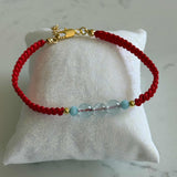 A red braided cord bracelet with a gold clasp and three light blue beads, displayed on a white fabric jewelry pillow against a light marble background.