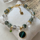 A beaded bracelet displayed on a white fabric cushion, featuring green and clear faceted beads, gold-tone chain and spacers, and a round green cat's eye pendant.