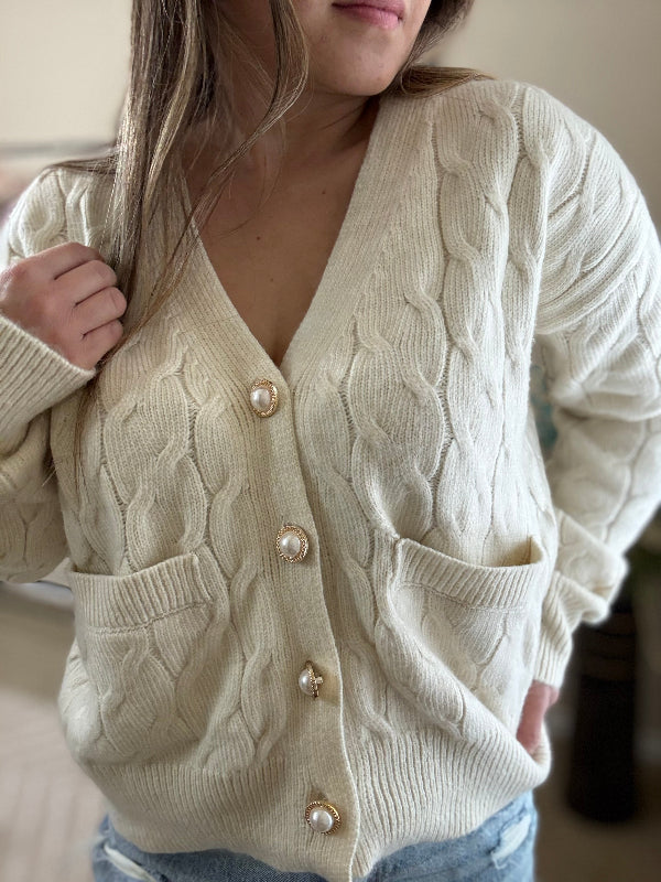 A beige cable knit button-up cardigan with front pockets and statement buttons, worn by a model against a casual background.