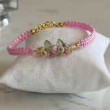 A pink braided cord bracelet with a gold clasp and a central floral charm, displayed on a white fabric jewelry stand over a light marble surface.