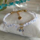 A delicate beaded bracelet with white and light blue beads, gold-toned chain hardware, a small gold heart charm, and two blue faceted beads, displayed on a white fabric cushion.