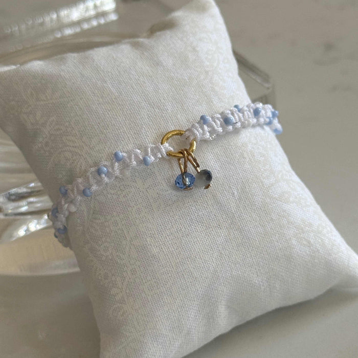 A delicate beaded bracelet with light blue and white beads, a gold charm ring, and a small blue stone charm, displayed on a white fabric pillow.