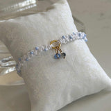 A delicate beaded bracelet with light blue and white beads, a gold charm ring, and a small blue stone charm, displayed on a white fabric pillow.