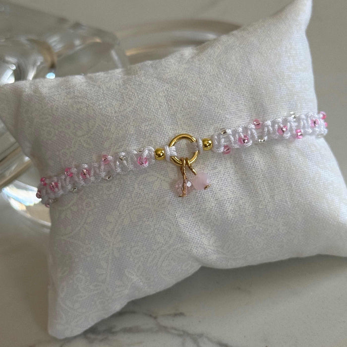 A delicate beaded bracelet with pink and clear beads, a gold ring clasp, and a small pink bow charm, displayed on a white fabric pillow over a marble surface.