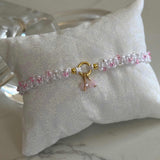 A delicate beaded bracelet with pink and clear beads, a gold ring clasp, and a small pink bow charm, displayed on a white fabric pillow over a marble surface.