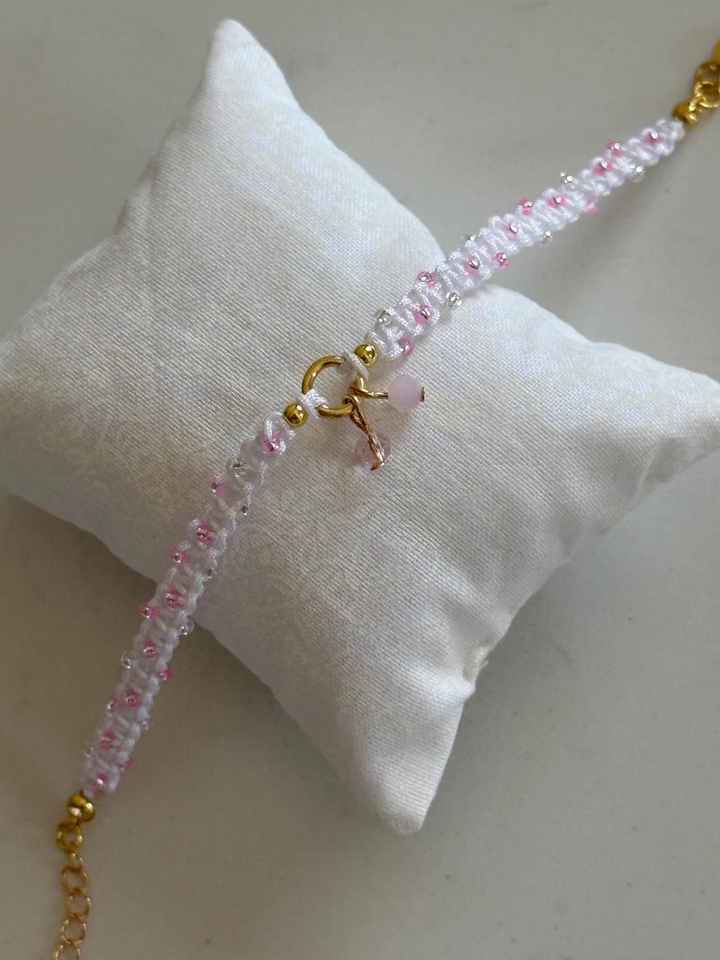 A delicate beaded bracelet with pink and clear beads, gold-toned metal links, and a small pink stone charm, displayed on a white fabric pillow.