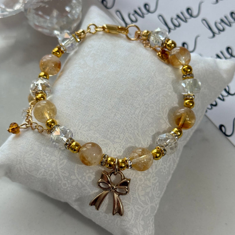A beaded bracelet with translucent amber and clear beads, gold-toned spacer beads, and a bronze bow charm, displayed on a white textured cushion.