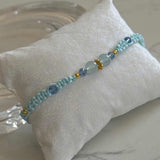 A light blue beaded bracelet with gold spacer beads and larger pale blue stone beads, displayed on a white fabric pillow over a light marble surface.