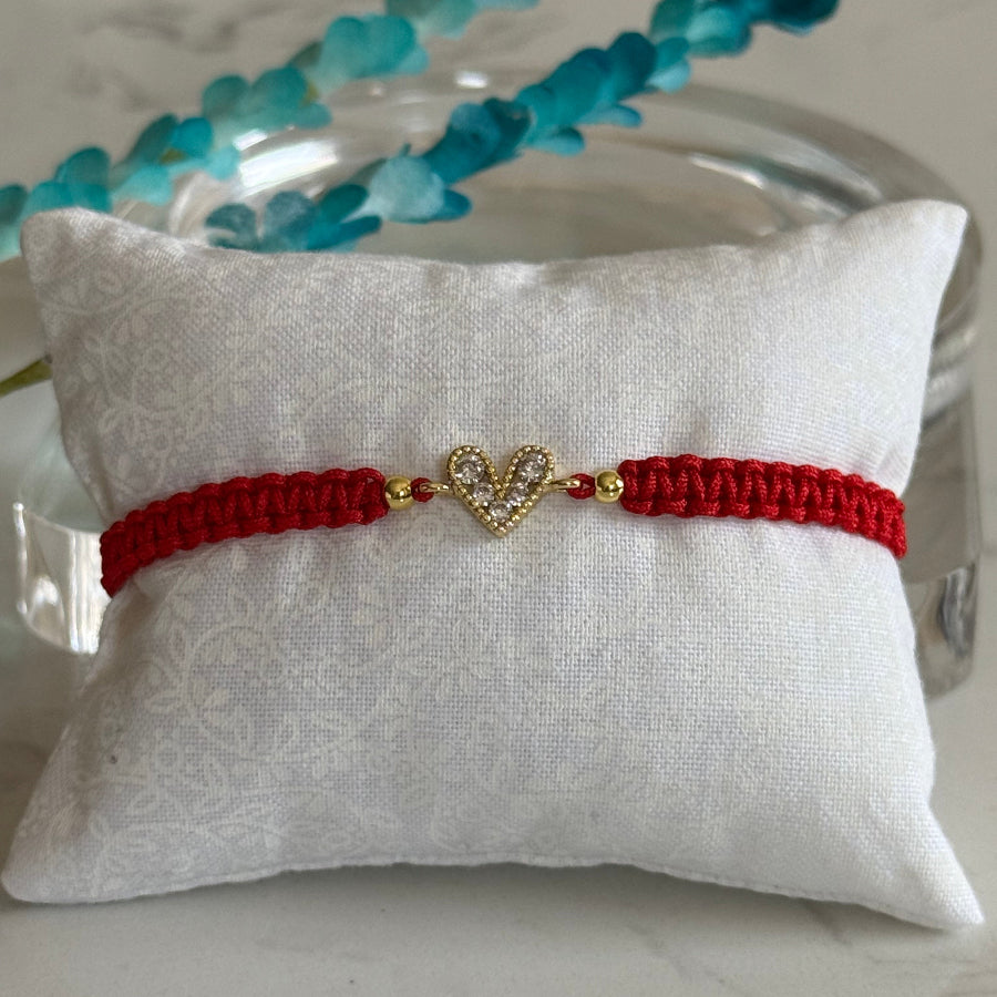 A red braided string bracelet with a gold heart-shaped charm set with small clear stones, displayed on a white textured pillow.