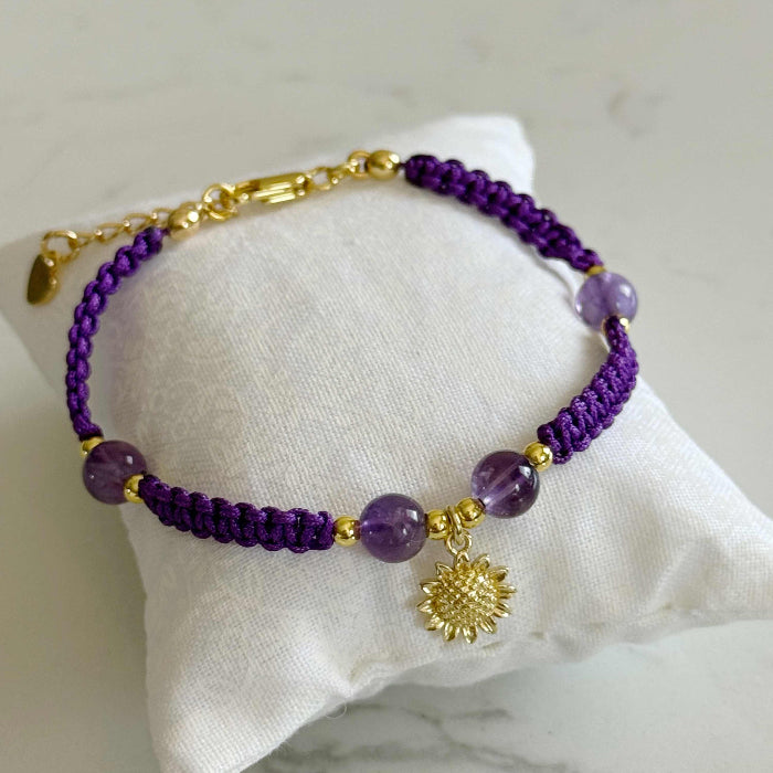 A purple beaded bracelet with gold-toned metal accents, including a sun charm and clasp, displayed on a white fabric pillow over a light marble surface.