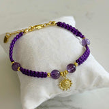 A purple beaded bracelet with gold-toned metal accents, including a sun charm and clasp, displayed on a white fabric pillow over a light marble surface.