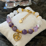 A beaded bracelet with purple amethyst and white pearl-like beads, gold-toned spacer beads, and a gold heart charm featuring an eye motif, displayed on a cream fabric cushion.