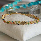 A delicate gold beaded bracelet with small multicolored beads and a central gold heart charm, displayed on a white fabric cushion.