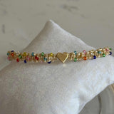 A delicate gold beaded bracelet with a central gold heart charm, displayed on a textured white fabric cushion.