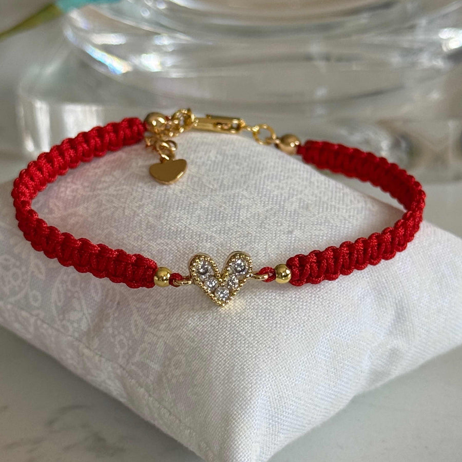 A red braided cord bracelet with gold-tone hardware, including a heart-shaped charm set with small clear stones, displayed on a white fabric cushion.