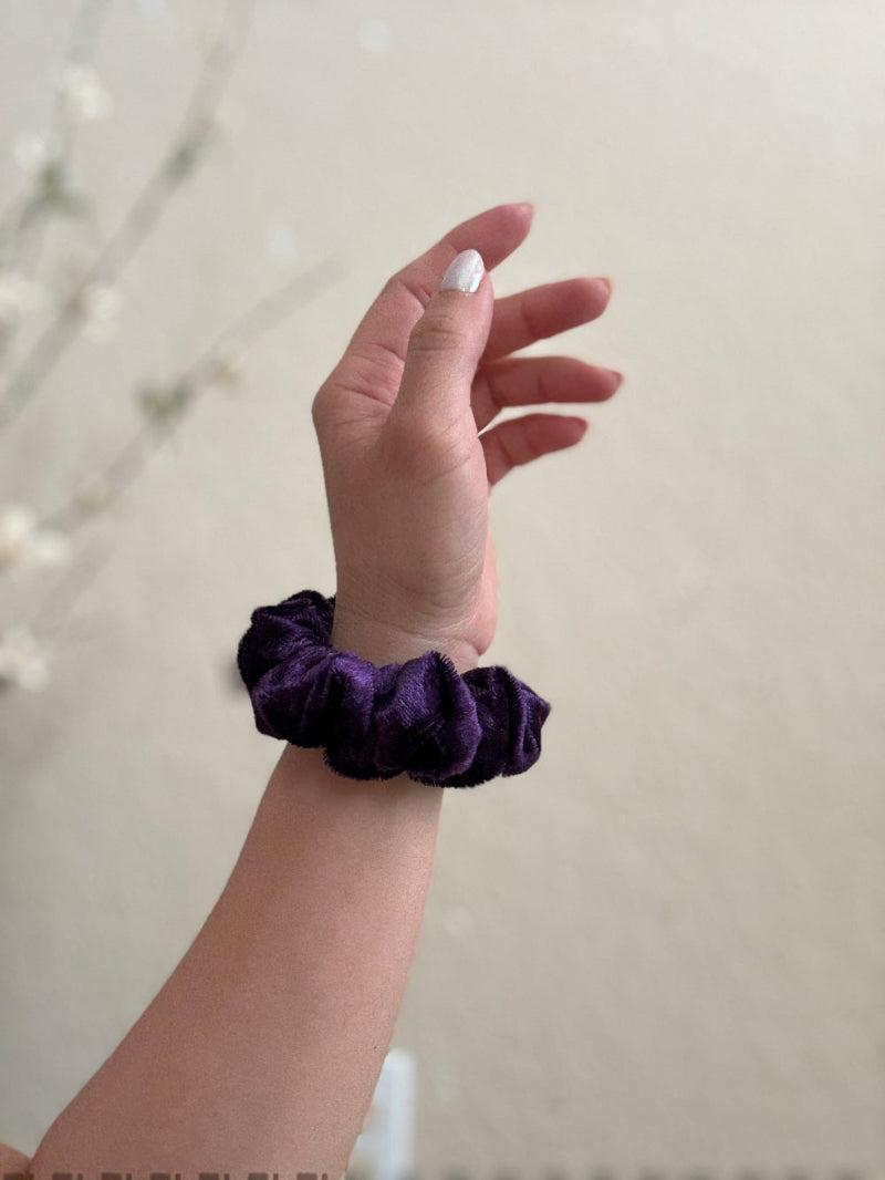 Hand wearing a purple scrunchie on a neutral background