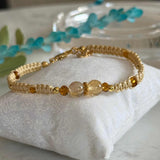 A gold beaded bracelet with a central clear faceted bead, displayed on a white textured fabric cushion.