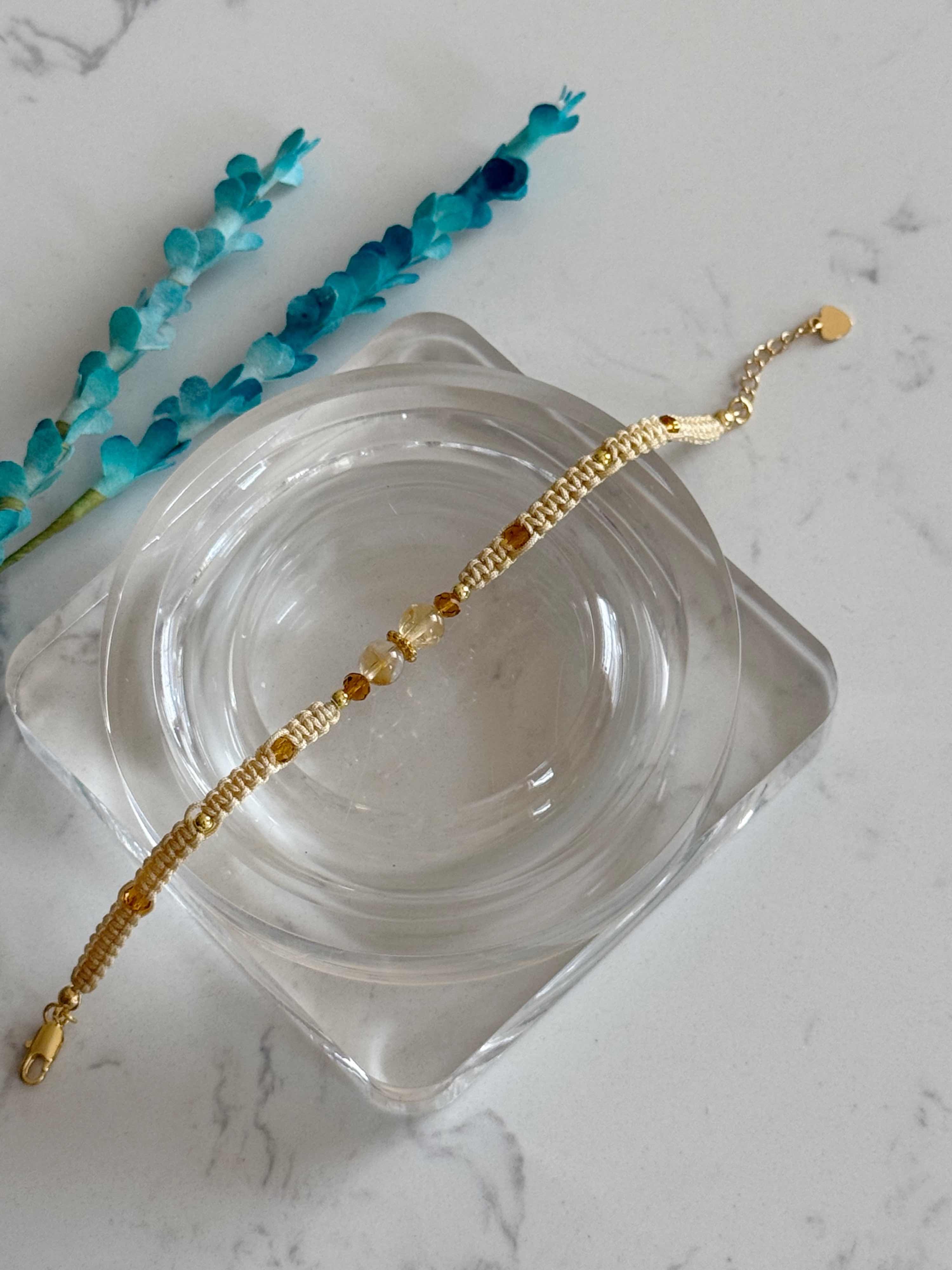 A delicate gold chain bracelet with small amber-colored beads, displayed on a clear glass dish over a white marble surface with blue decorative sprigs.