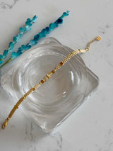 A delicate gold chain bracelet with small amber-colored beads, displayed on a clear glass dish over a white marble surface with blue decorative sprigs.