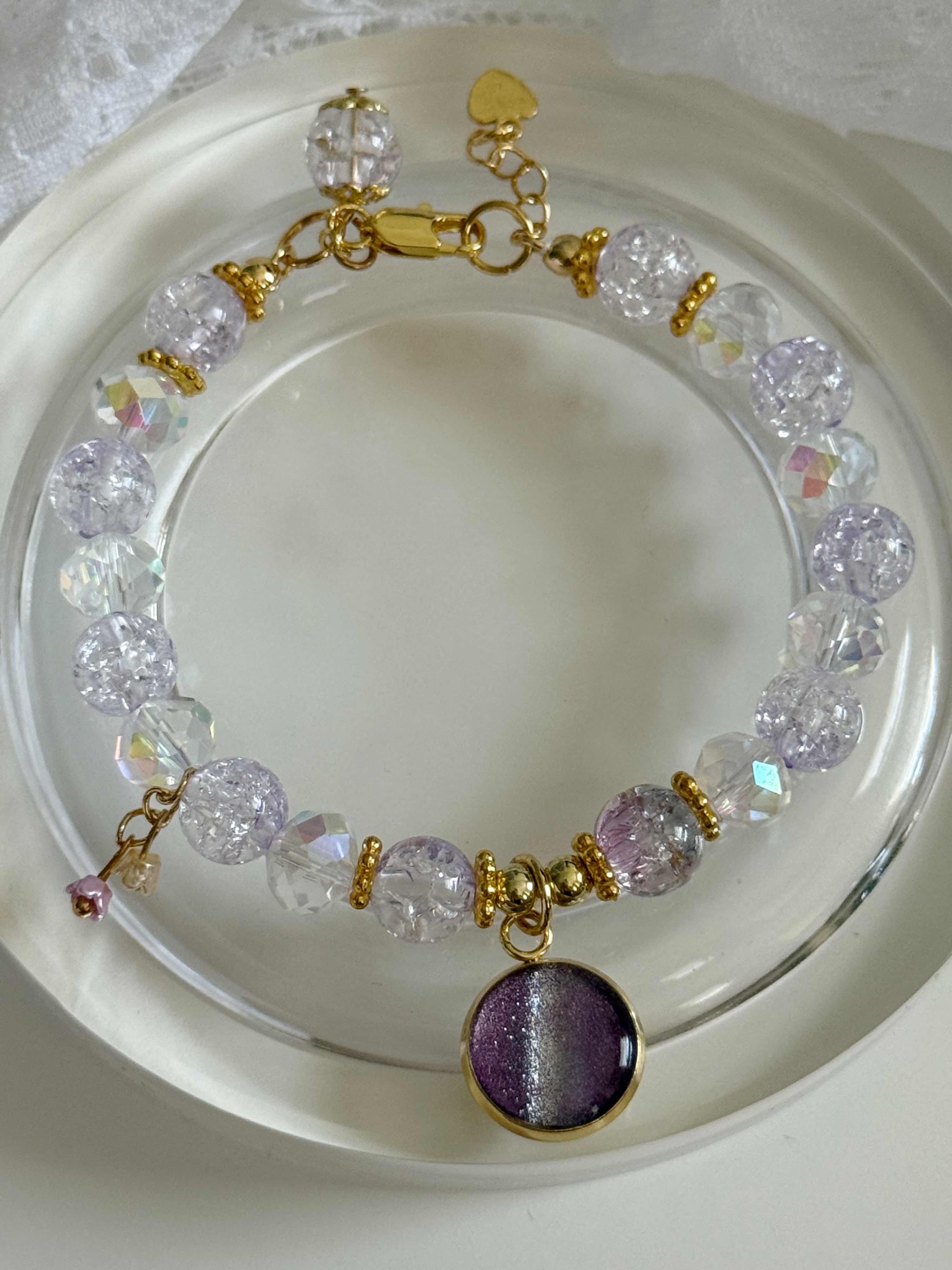 A beaded bracelet with clear faceted beads, gold-tone spacer rings, and a purple glitter oval pendant, displayed on a white dish.