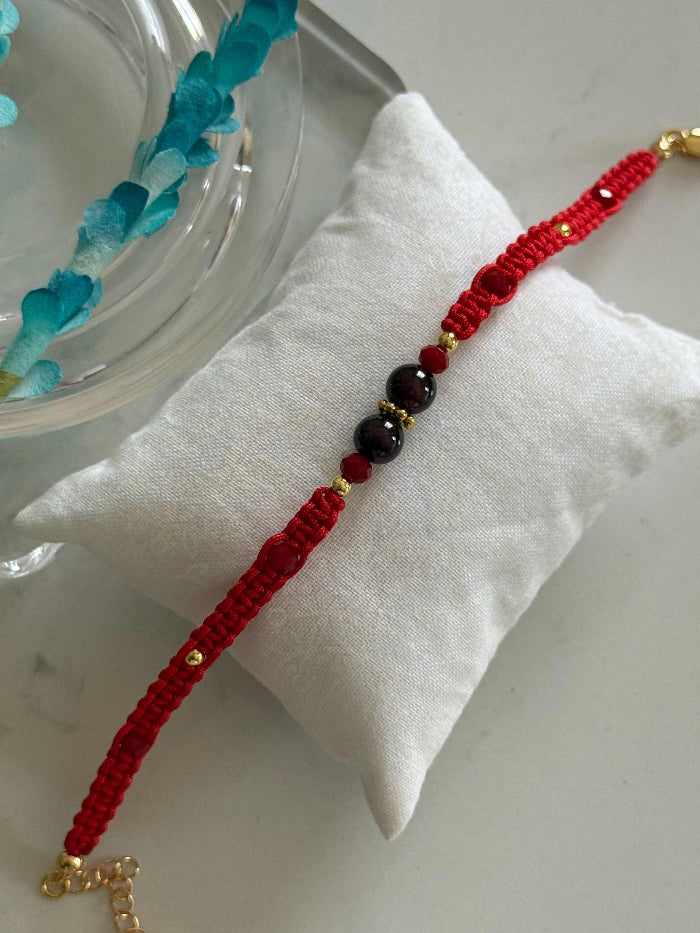 A red braided cord bracelet with dark red beads and small gold-toned accents, displayed on a white cushion.
