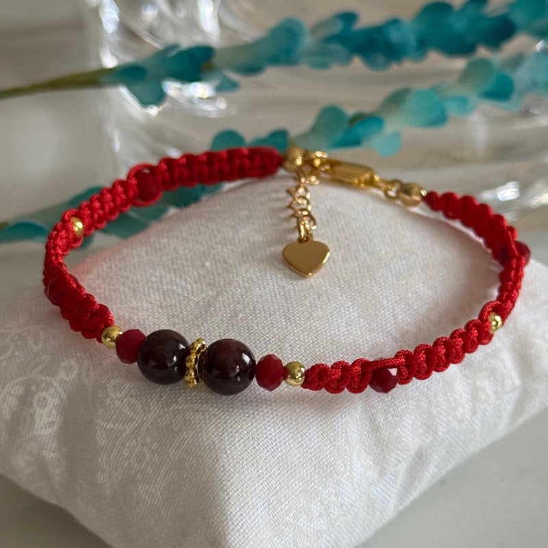 A red braided cord bracelet with dark red beads, gold spacer details, and a small gold heart charm, displayed on a white fabric cushion.