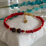 A red braided cord bracelet with dark red beads, gold spacer details, and a small gold heart charm, displayed on a white fabric cushion.