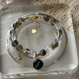 A beaded charm bracelet displayed on a clear acrylic tray. The bracelet features dark green and clear faceted beads, gold-toned metal links and spacer beads, and a prominent round black and teal striped snake-eye style charm.