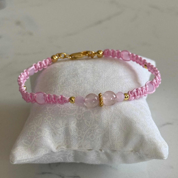 A pink beaded bracelet with gold-toned metal accents and a central light pink stone bead, displayed on a white fabric jewelry cushion over a light marble surface.