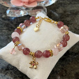 A pink and rose quartz beaded bracelet with gold-toned hardware and a small flower charm, displayed on a cream fabric cushion.
