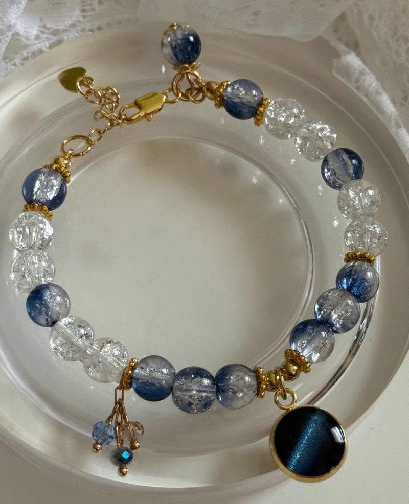 A beaded bracelet with alternating clear faceted beads and deep blue marbled beads, accented by gold-tone links and a round dark blue cat-eye charm, displayed on a white plate.